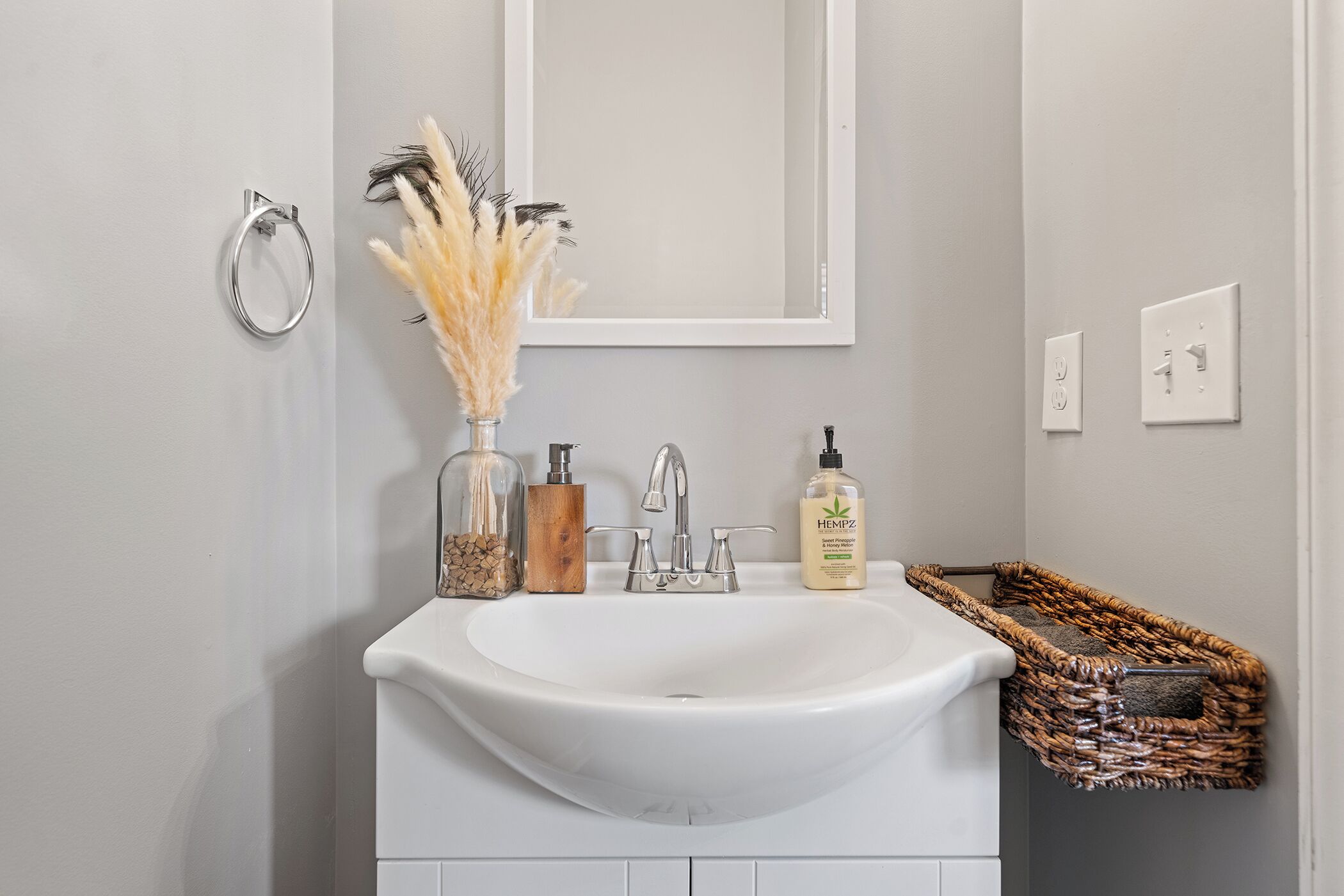 Une salle de bain avec un lavabo blanc, un miroir, un porte-serviettes, une bouteille de shampoing et un panier en osier.