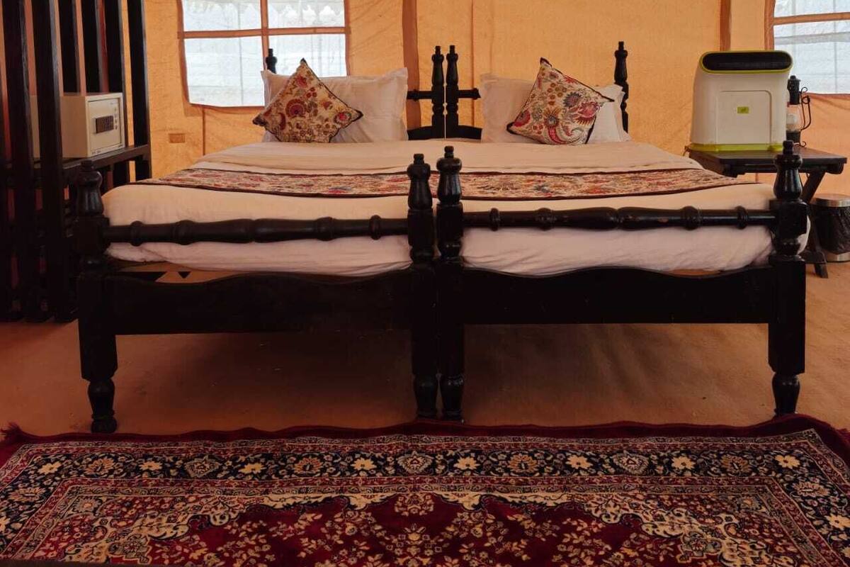 Un lit traditionnel avec un cadre en bois, deux oreillers et une couvre-lit à motifs. Il y a une fenêtre avec un rideau léger et une petite table avec une lampe placée à côté du lit.