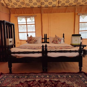 Un lit traditionnel avec un cadre en bois, deux oreillers et une couvre-lit à motifs. Il y a une fenêtre avec un rideau léger et une petite table avec une lampe placée à côté du lit.