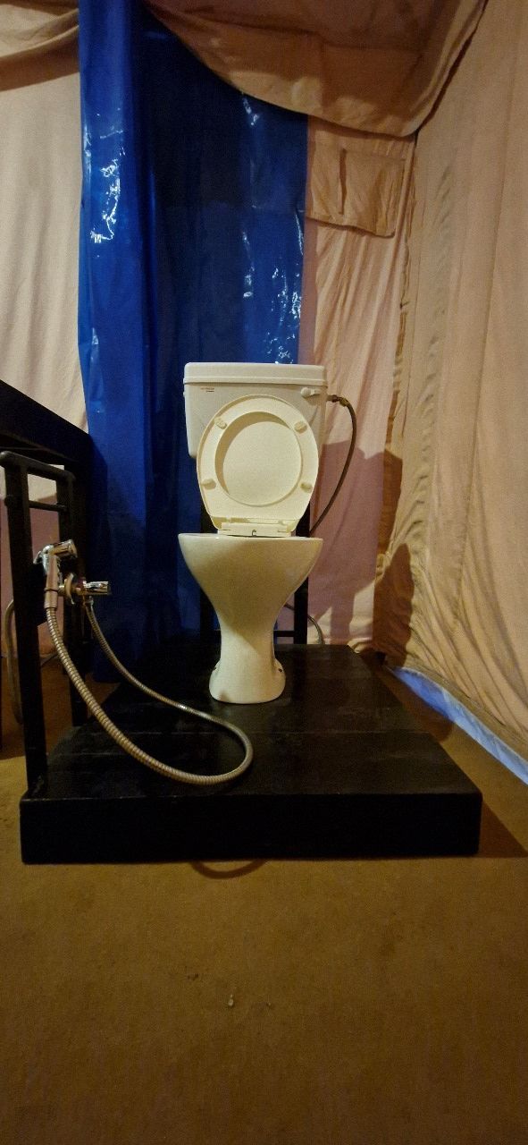Des toilettes équipées d’un tuyau d’arrosage, installées sur une plateforme noire à l’intérieur d’une structure ressemblant à une tente.
