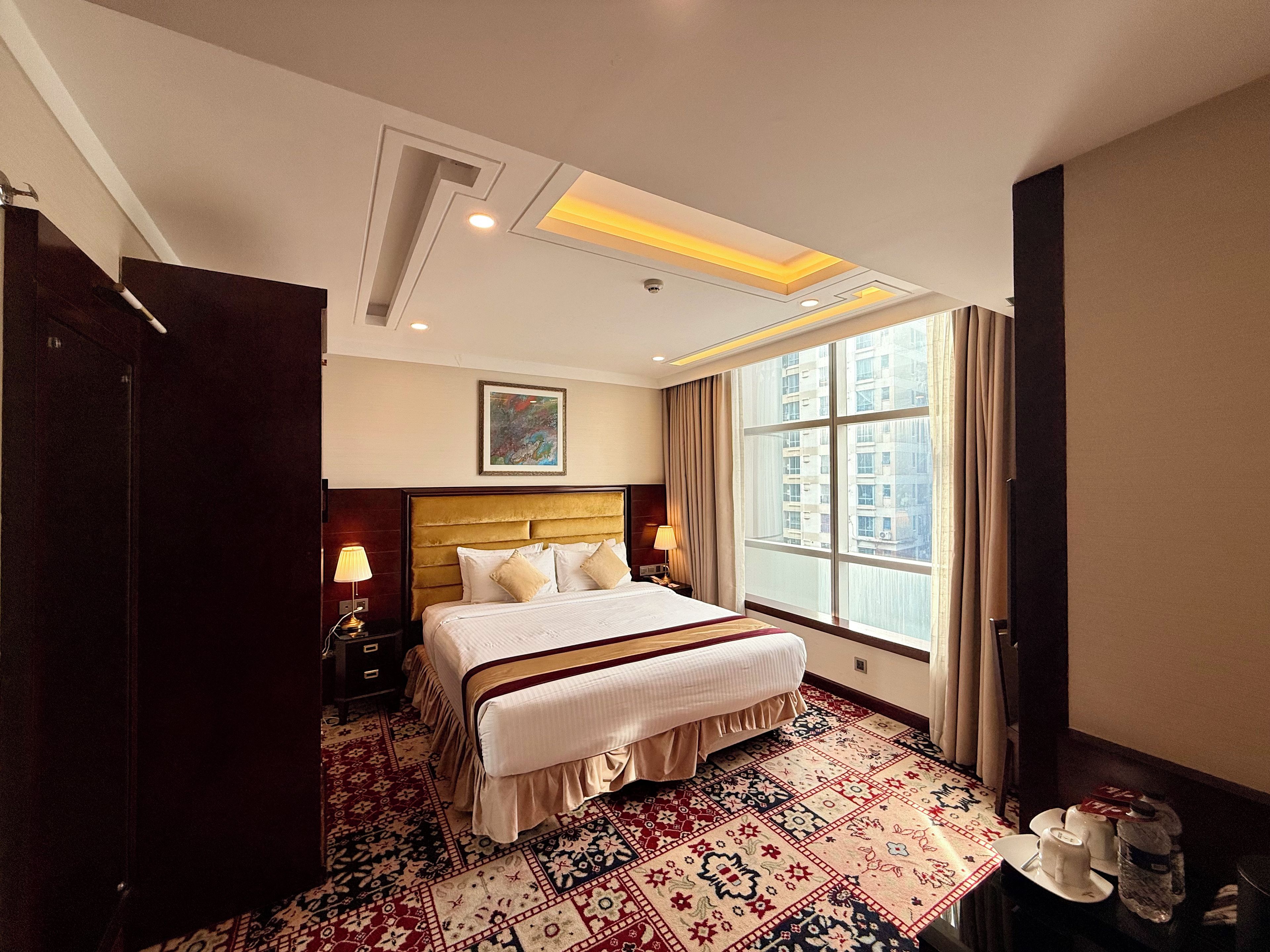 Quarto de hotel com uma cama grande, janela com cortinas, tapete estampado e mesa de cabeceira com abajur.