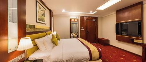A hotel room with a large bed, a bedside table with a lamp, a telephone, a TV, and a door leading to a bathroom.