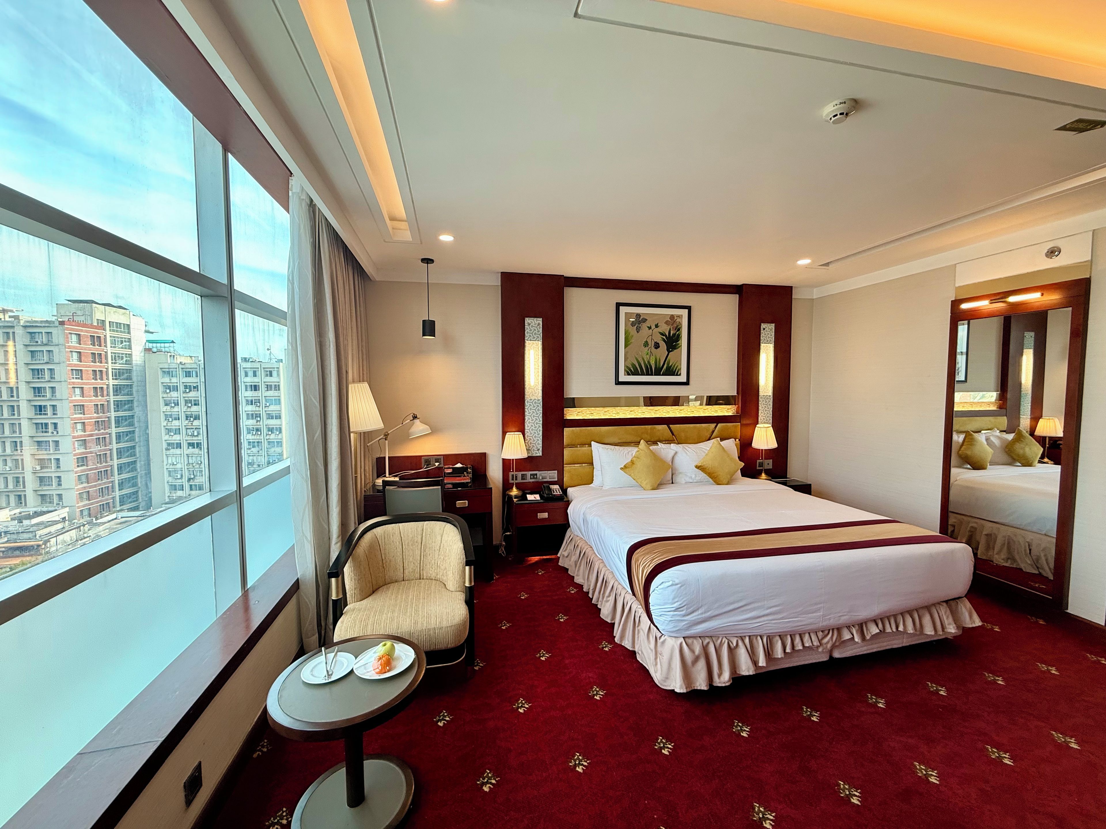 A hotel room with a large bed, a desk, a chair, a small table with plates, and a large window offering a city view.