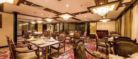 Un amplio salón de banquetes con varias mesas dispuestas para comer, candelabros ornamentados y un suelo cubierto de alfombra tradicional.