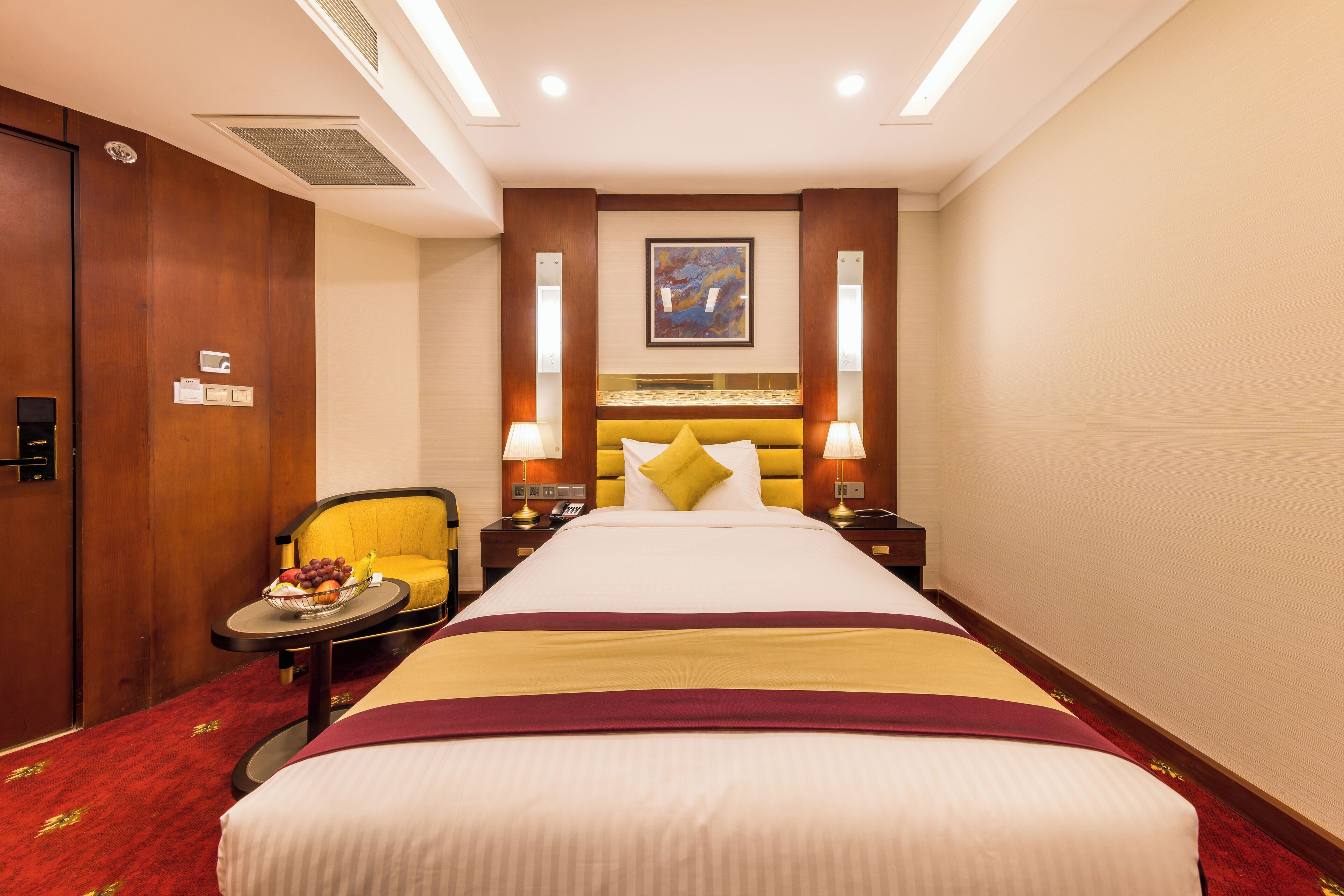 A hotel room with a large bed, a bedside table with a fruit bowl, a chair, and two bedside lamps.