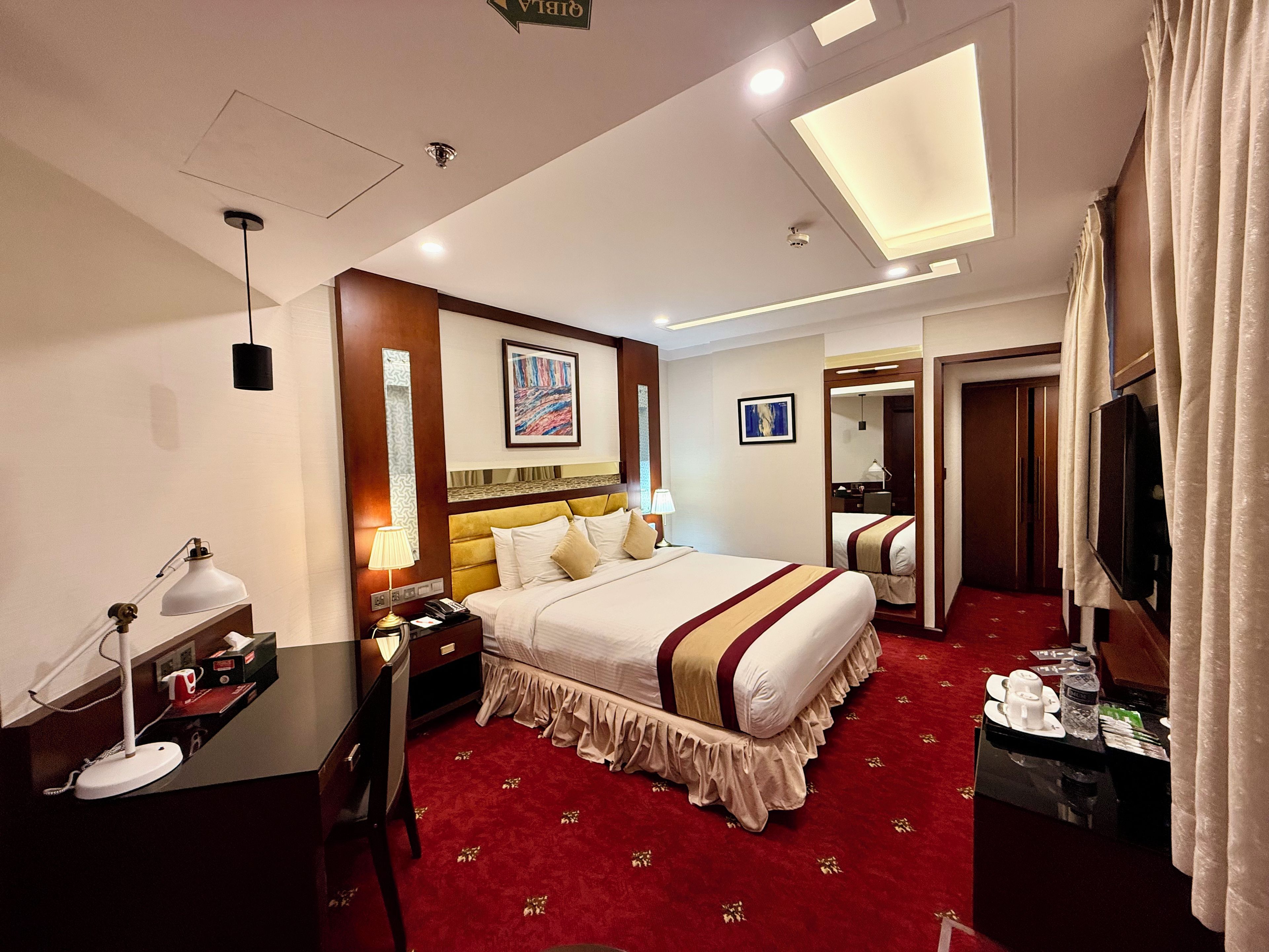 A hotel room with a large bed, bedside tables, a desk, a chair, a television, and a mirror.