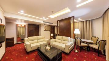A hotel room with two beige sofas, a glass coffee table, a small round table with chairs, a lamp, and a red carpet with a floral pattern.