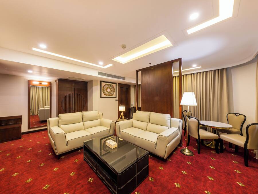 A hotel room with two beige sofas, a glass coffee table, a small round table with chairs, a lamp, and a red carpet with a floral pattern.