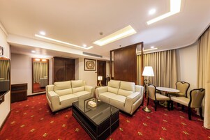 Habitación de hotel con dos sofás color beige, una mesa de centro de vidrio, una mesita redonda con sillas, una lámpara y una alfombra roja con estampado floral.