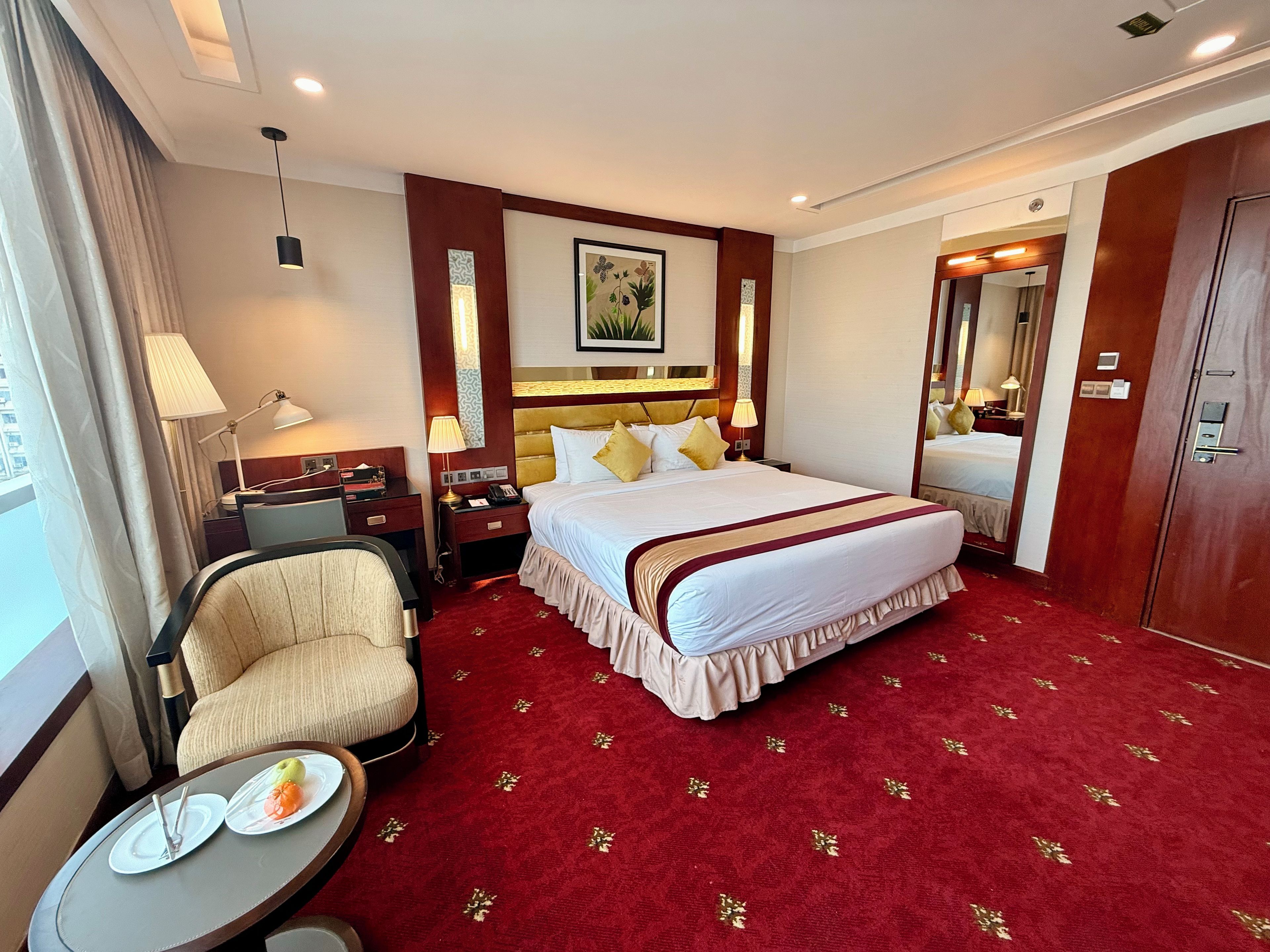 A hotel room with a large bed, a chair, a small table with plates, and a mirror.