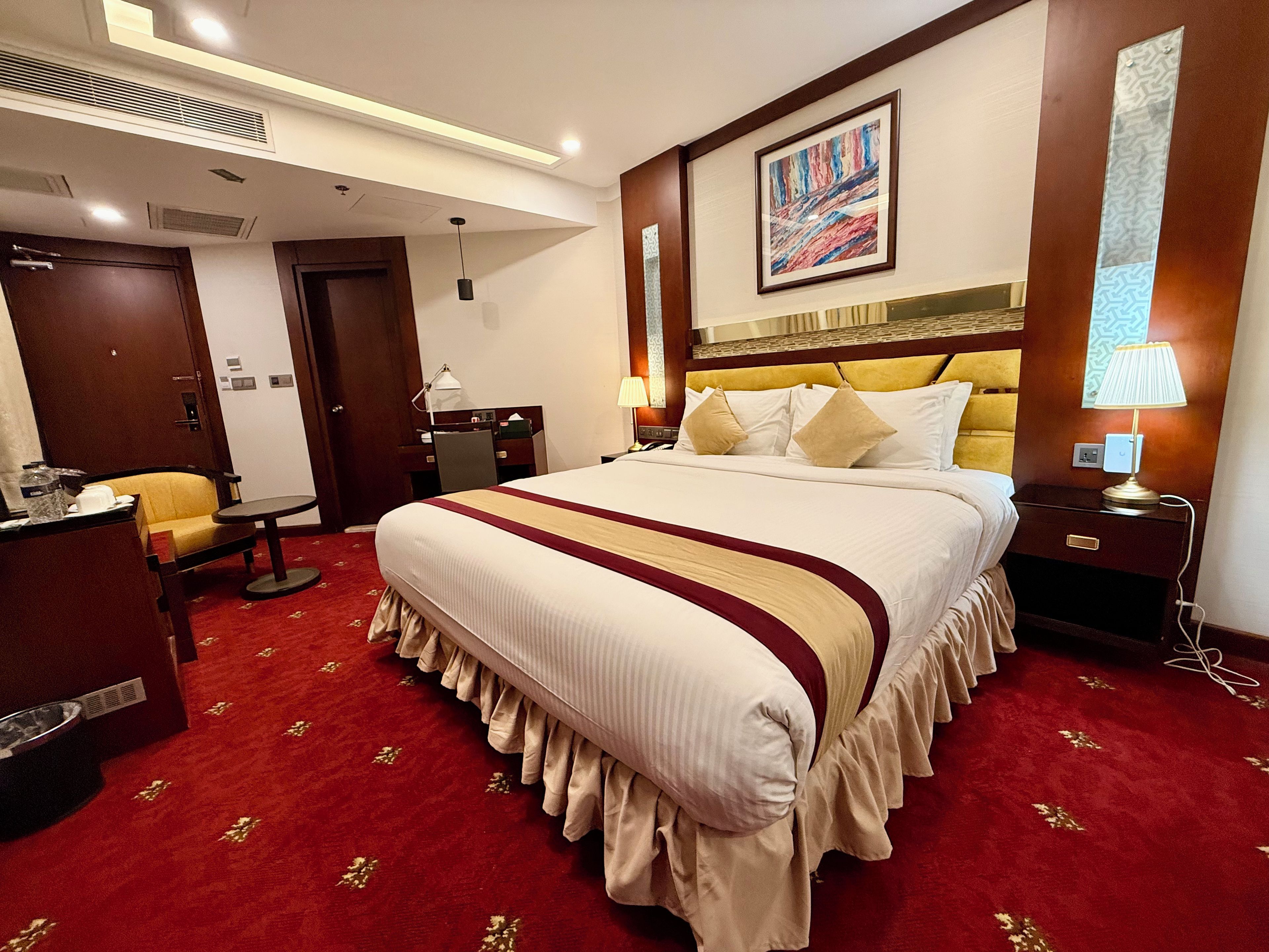 Quarto de hotel com cama grande, escrivaninha, cadeira, abajur e um quadro na parede.