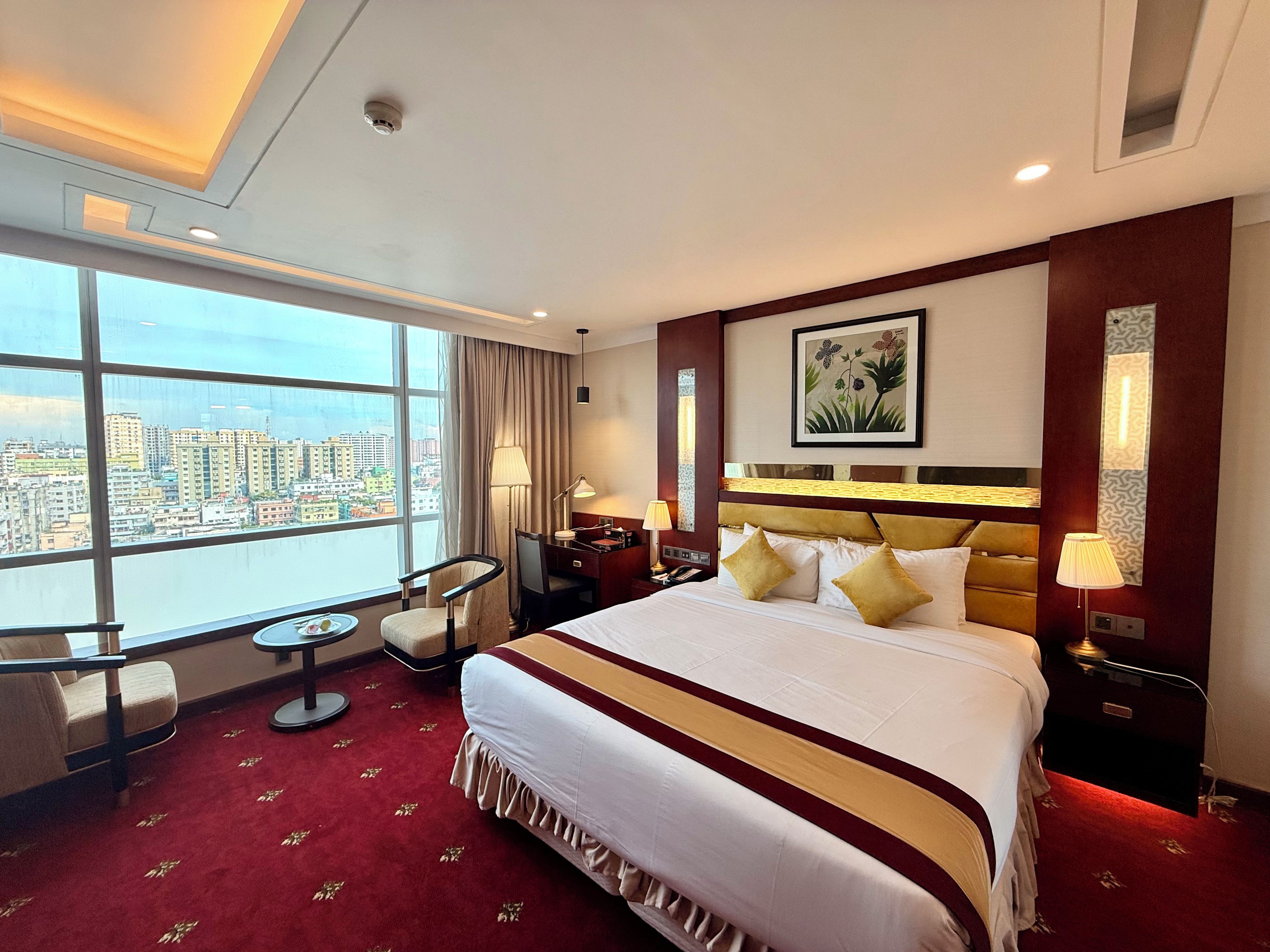 Ein Hotelzimmer mit einem großen Bett, einem Schreibtisch, einem Sessel, einem Nachttisch, einer Lampe und Blick auf die Stadt.
