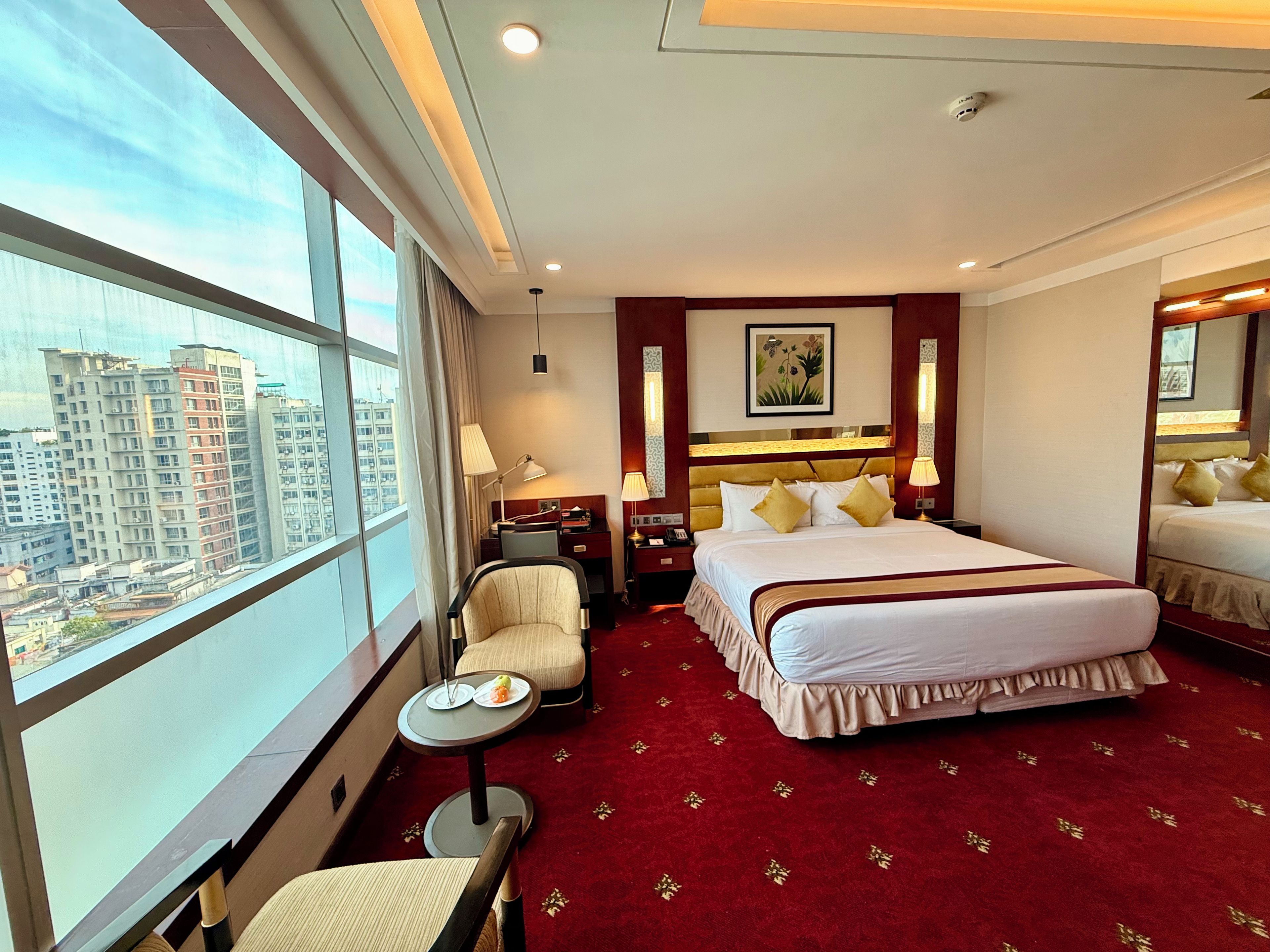 A hotel room with a large window, a bed with white bedding, a desk with a lamp, and a chair.