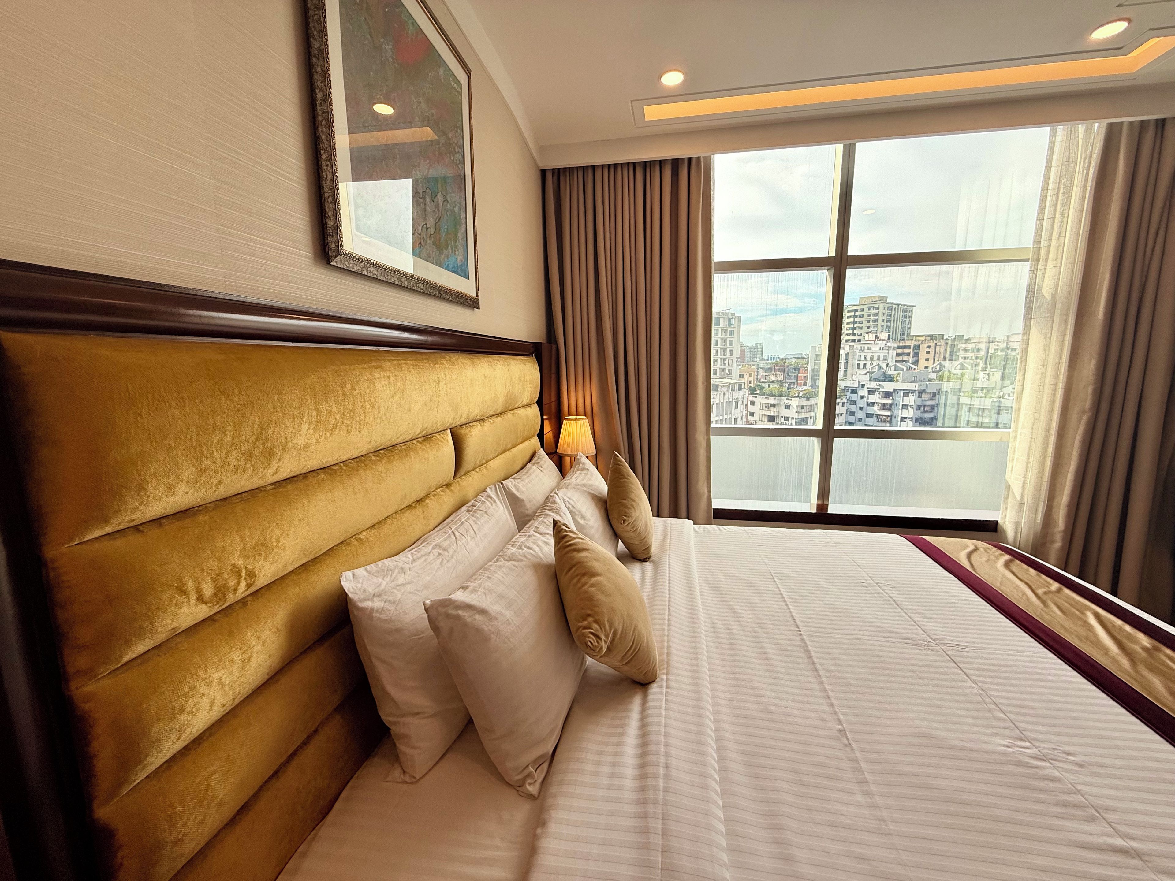 Ein Hotelzimmer mit einem großen Bett, einem Kopfteil, einem Fenster mit Vorhängen und Blick auf die Stadt.