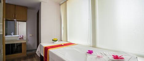 A single bed with a white bedspread and a red and yellow towel, a small table with a vase of flowers, and a window with curtains.