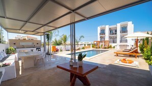 Un espacio exterior techado con piscina, sillas de descanso, una mesa y vista a un edificio blanco.