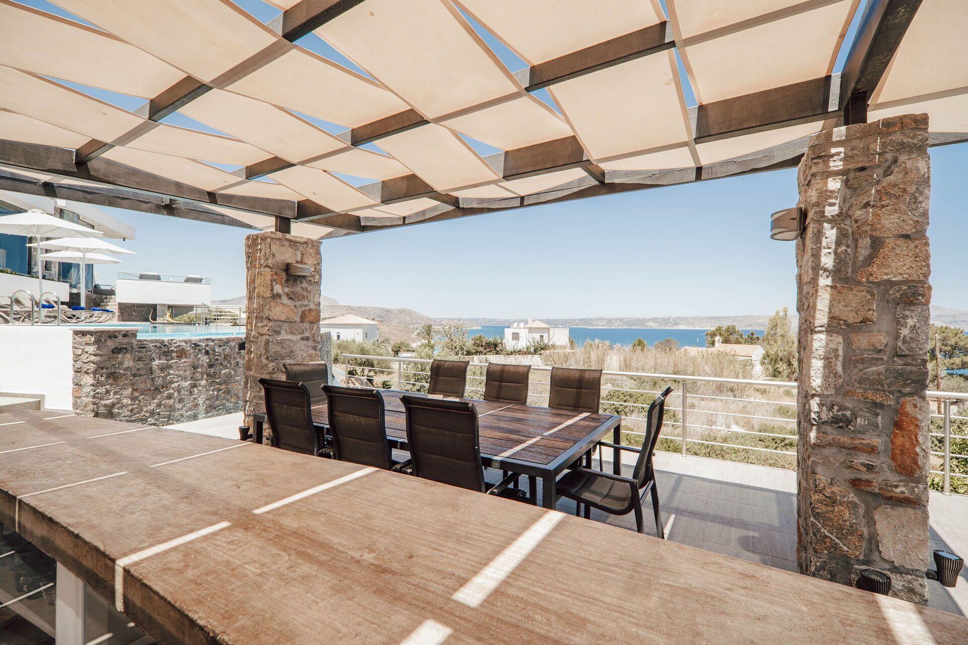 Un espace extérieur aménagé pour les repas, avec une colonne en pierre, une table en bois et des chaises, offrant une vue panoramique sur une piscine et des bâtiments situés au loin.