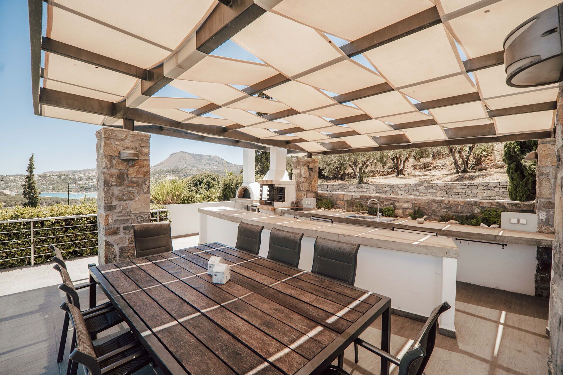 An outdoor kitchen area with a stone wall, a wooden table, and a pergola overhead.