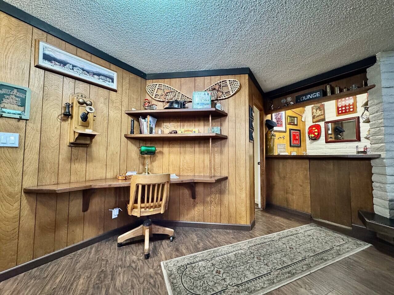 Habitación de esquina con paredes de madera, un escritorio con silla, estanterías con diversos objetos y una pequeña zona de bar con un cartel.