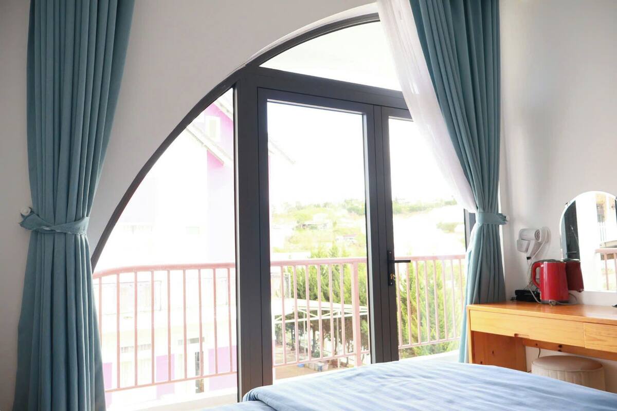A bedroom with a bed, a desk, a chair, a window with curtains, and a balcony with a railing.