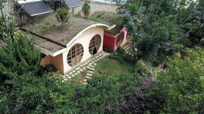 Ein kleines, eingeschossiges Gebäude mit einem geneigten Dach, einer roten Tür und einem Garten mit verschiedenen Pflanzen und einem Weg.