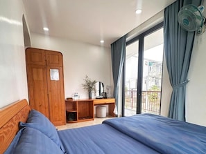 A bedroom with a bed, wooden wardrobe, desk with a mirror, and a balcony with a view of buildings. - Otel Dalat (Xuan Truong - Da Lat)