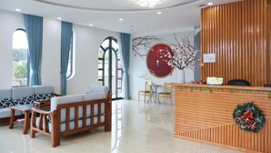 Un hall d’hôtel avec un comptoir d’accueil en bois, un espace salon avec des canapés et des fauteuils, et de grandes fenêtres ornées de rideaux bleus.