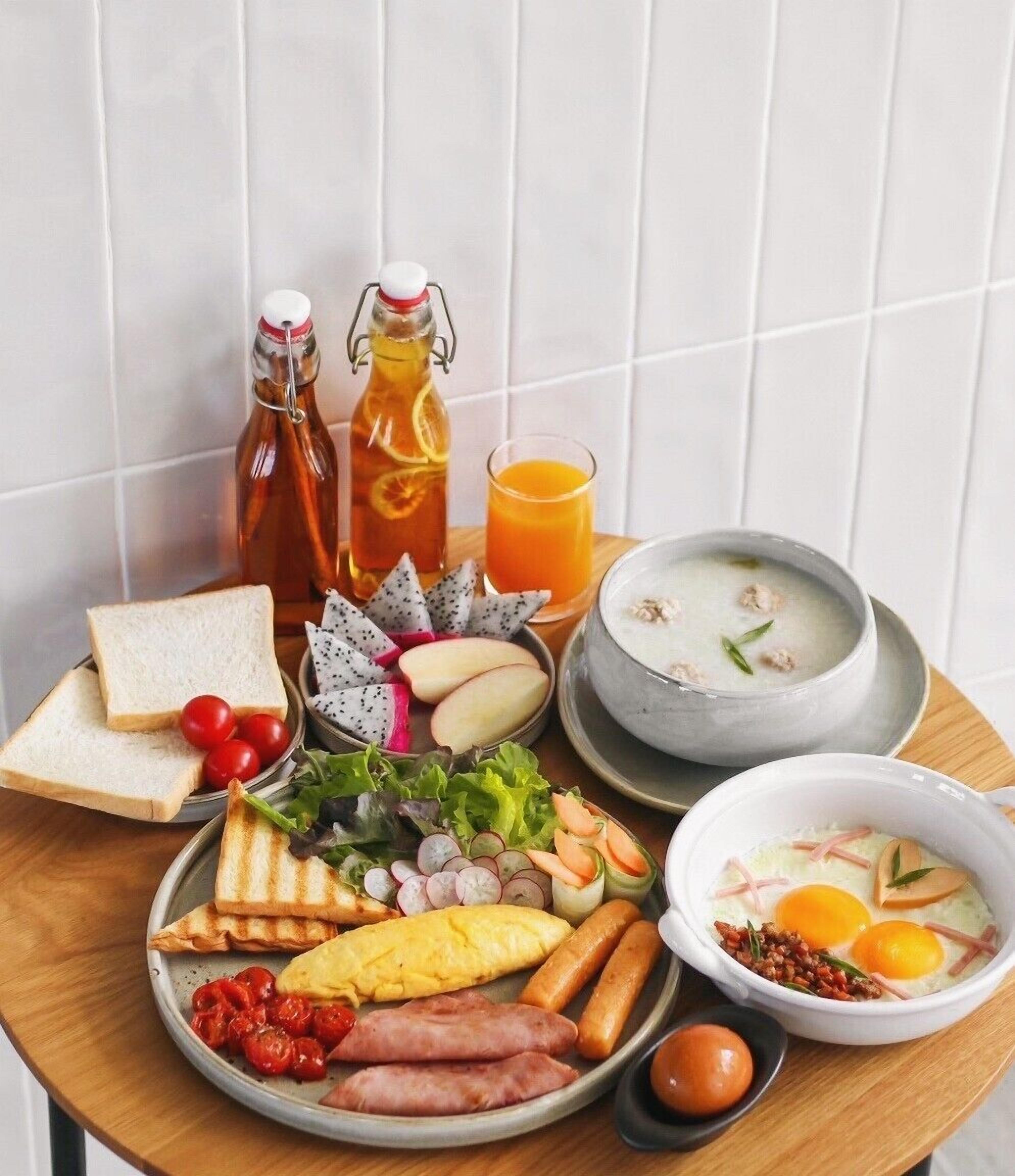 A breakfast spread on a wooden table including toast, eggs, sausages, and a bowl of soup.