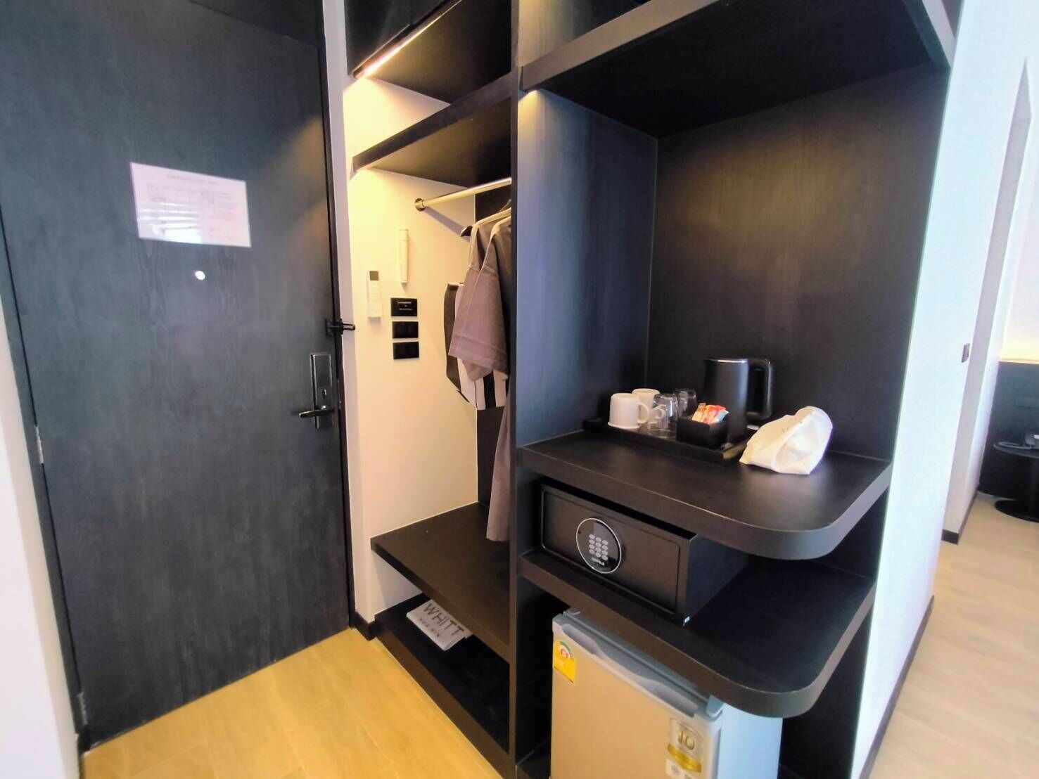 A compact hotel room with a dark wardrobe, a small desk with a coffee maker, and a mini fridge.