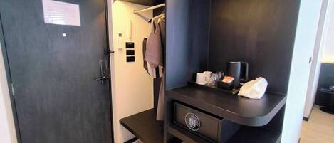 A compact hotel room with a dark wardrobe, a small desk with a coffee maker, and a mini fridge.