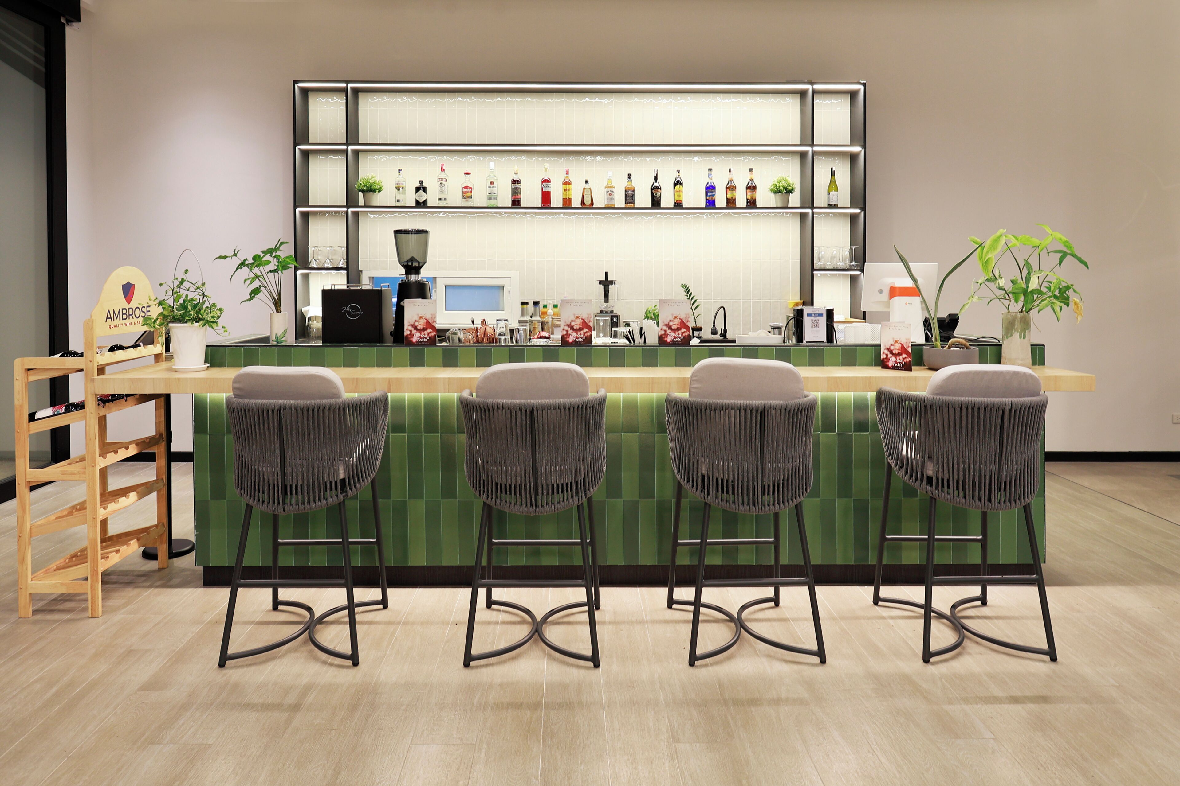 A bar area with a green tiled counter, four wicker bar stools, a wooden shelf with bottles, and a small plant.