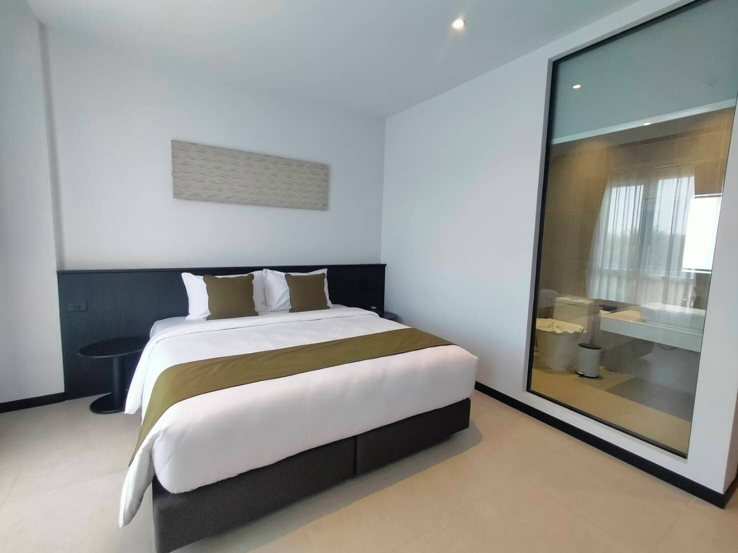 A modern hotel room with a large bed, a bedside table, and a glass partition separating the room from the bathroom.