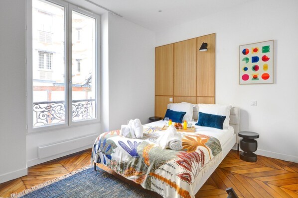 A modern bedroom with a large bed, a wooden wardrobe, a window with a view of buildings, and a colorful abstract painting.