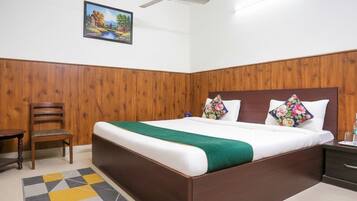 Un dormitorio con cabecera de madera, cama con ropa blanca y un plaid verde, mesita de noche de madera con lámpara, y una silla.