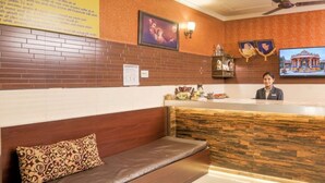 A reception area with a wooden reception desk, a cushioned bench, and a wall-mounted television displaying a building. - Shri Sai Guest House - Opp Azadpur Main Mandi (New Delhi)