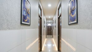 A hallway with white tiled walls and floor, framed artwork on the walls, and a door at the end. - Shri Sai Guest House - Opp Azadpur Main Mandi (New Delhi)