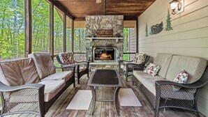 A covered outdoor seating area with wicker furniture, a stone fireplace, and a view of the forest.