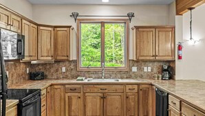 A kitchen with wooden cabinets, a stainless steel refrigerator, a double sink, and a window with a view of greenery.