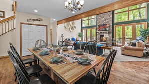 A dining area with a wooden table set for six, surrounded by chairs. A stone fireplace with a neon sign is visible in the background.