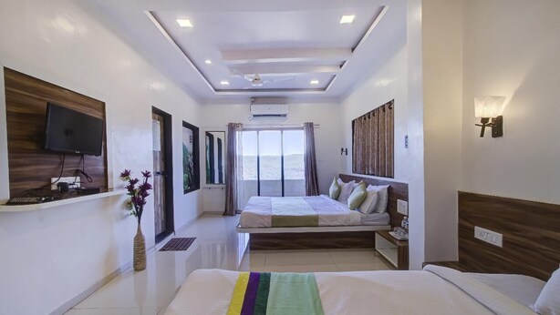 A modern hotel room with a large bed, a desk with a computer, a flat-screen TV mounted on the wall, and a large window providing a view of the sea. - Straw Bella Inn Villa (Dhangarwadi)