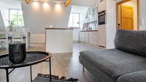 Una sala de estar moderna con un sofá gris, una mesa redonda con un objeto negro y una cocina al fondo.