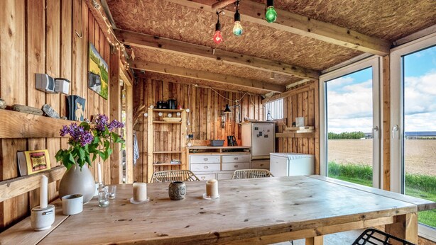 Un interior rústico de madera con una gran mesa de comedor, sillas, una zona de cocina y ventanas amplias que ofrecen vistas al exterior.