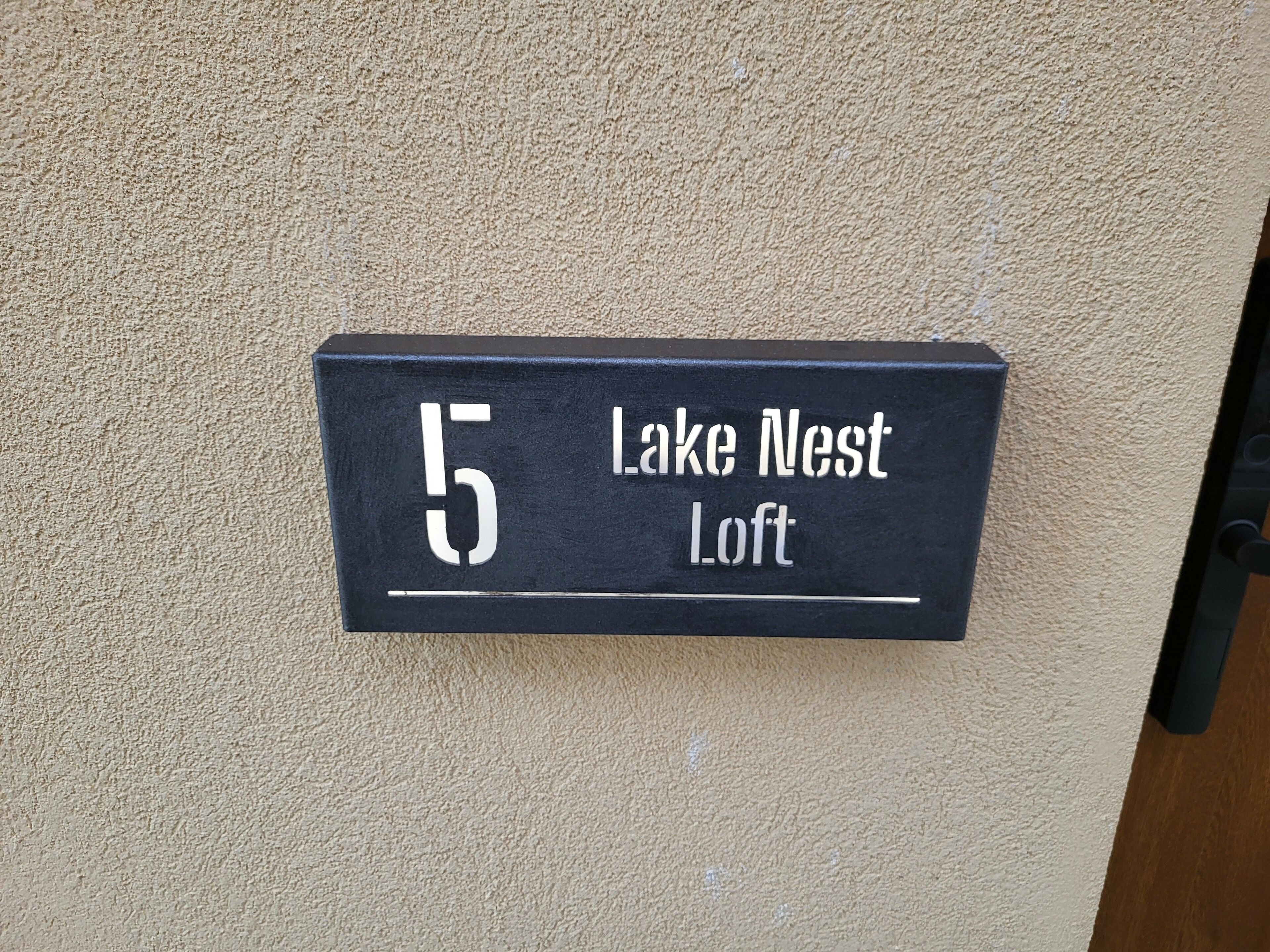A black house number sign with the text "5 Lake Nest Loft" mounted on a beige wall.