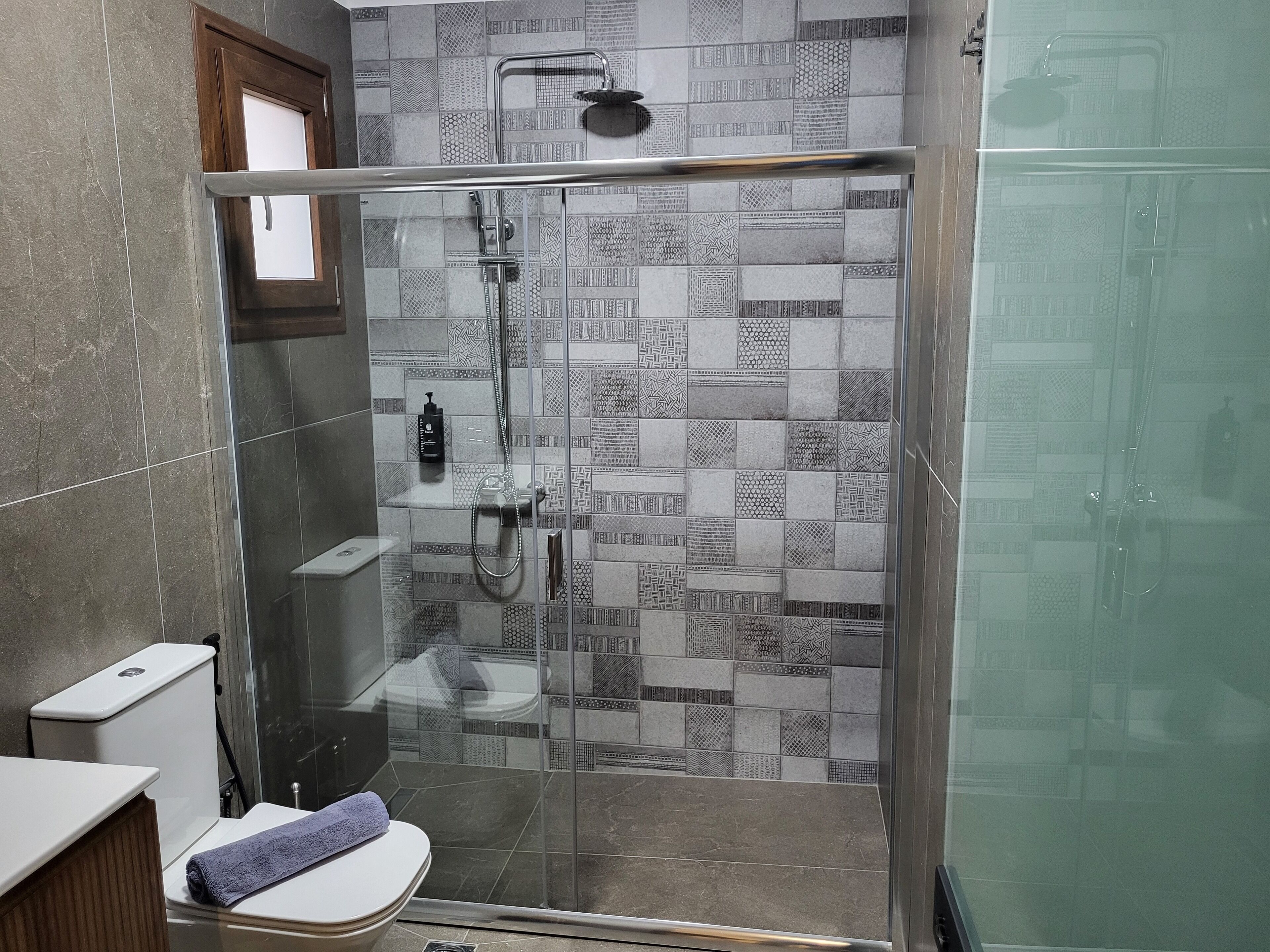 Un baño moderno con ducha de vidrio, baldosas estampadas, inodoro y lavamanos.