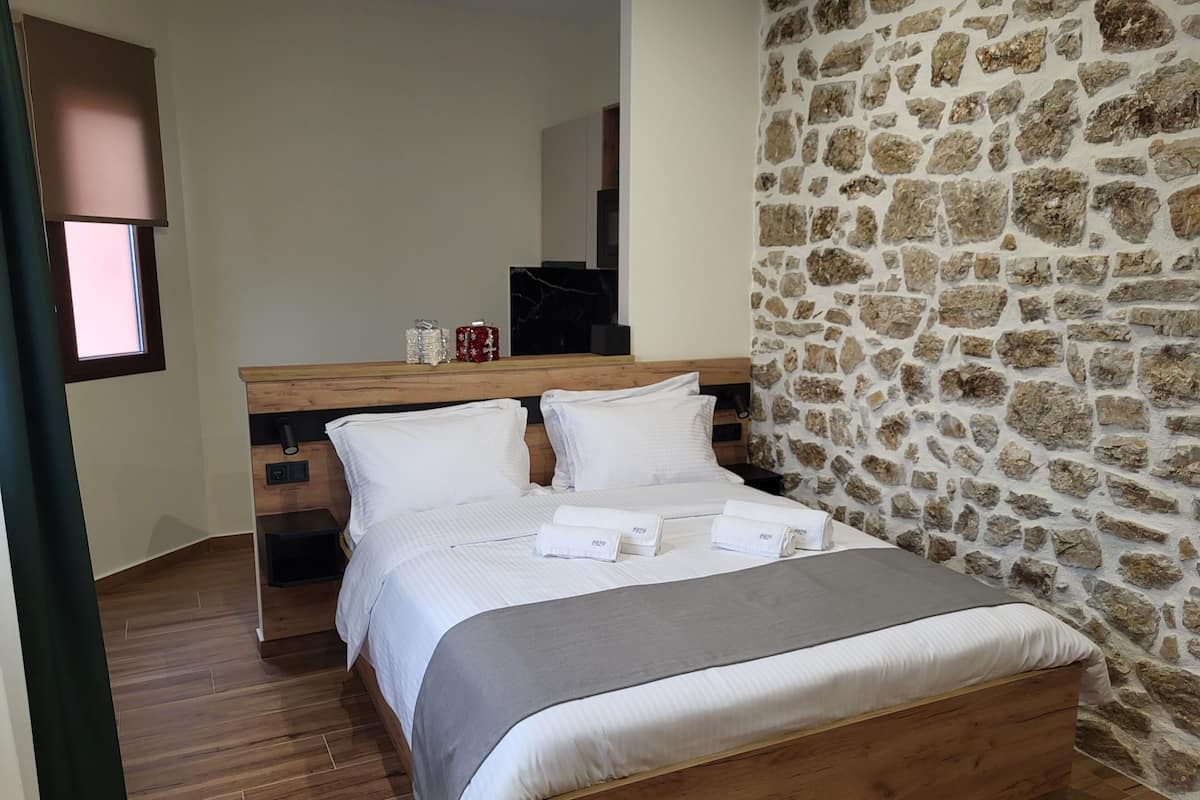 Une chambre avec un lit en bois, du linge de lit blanc, un mur en pierre et une fenêtre avec des stores.