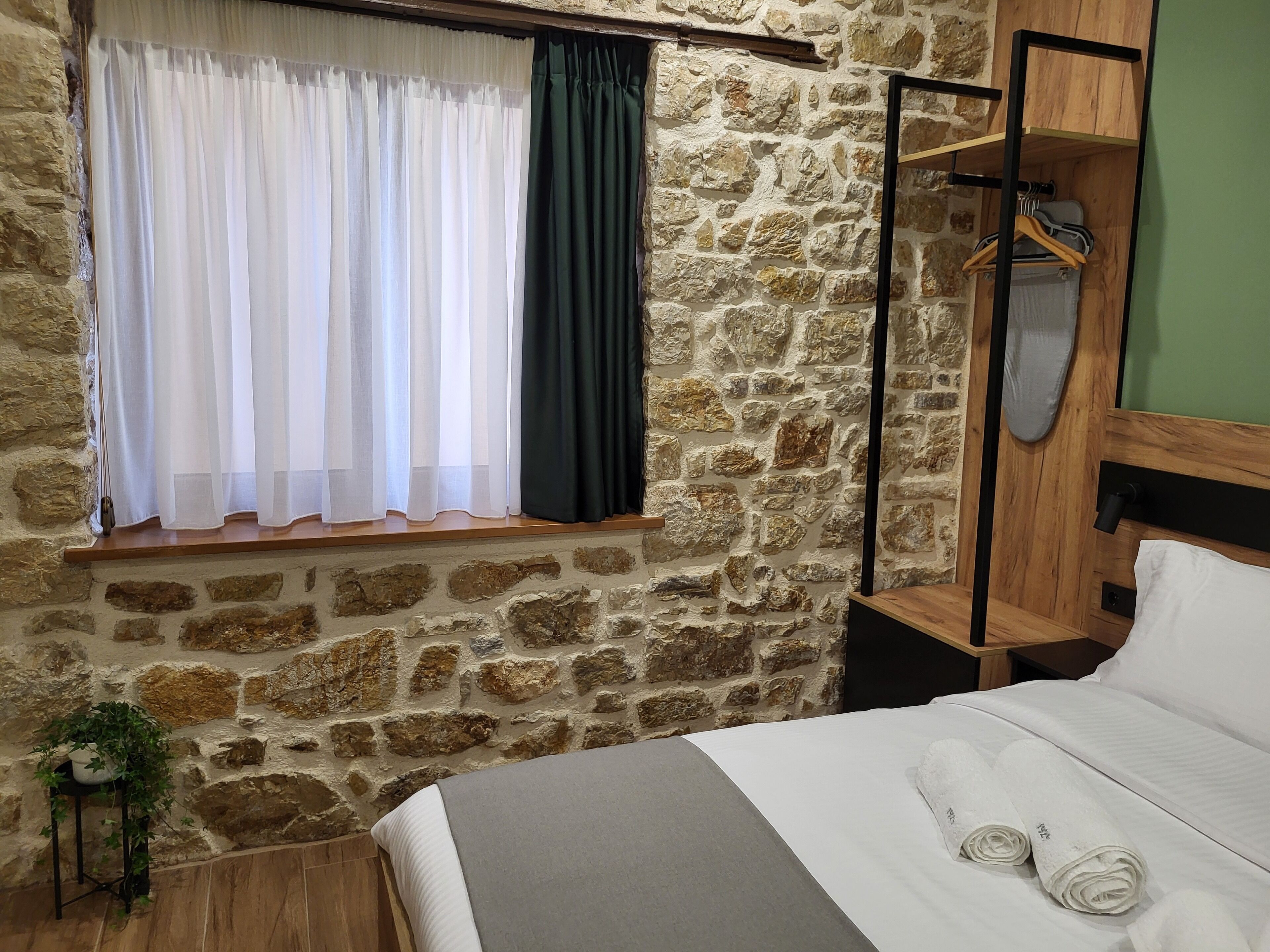 Quarto com parede de pedra, janela com cortinas brancas e verde escuro, estrutura de cama de madeira e mesa de cabeceira de madeira com espelho e cabide.