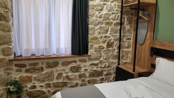 Quarto com parede de pedra, janela com cortinas brancas e verde escuro, estrutura de cama de madeira e mesa de cabeceira de madeira com espelho e cabide.