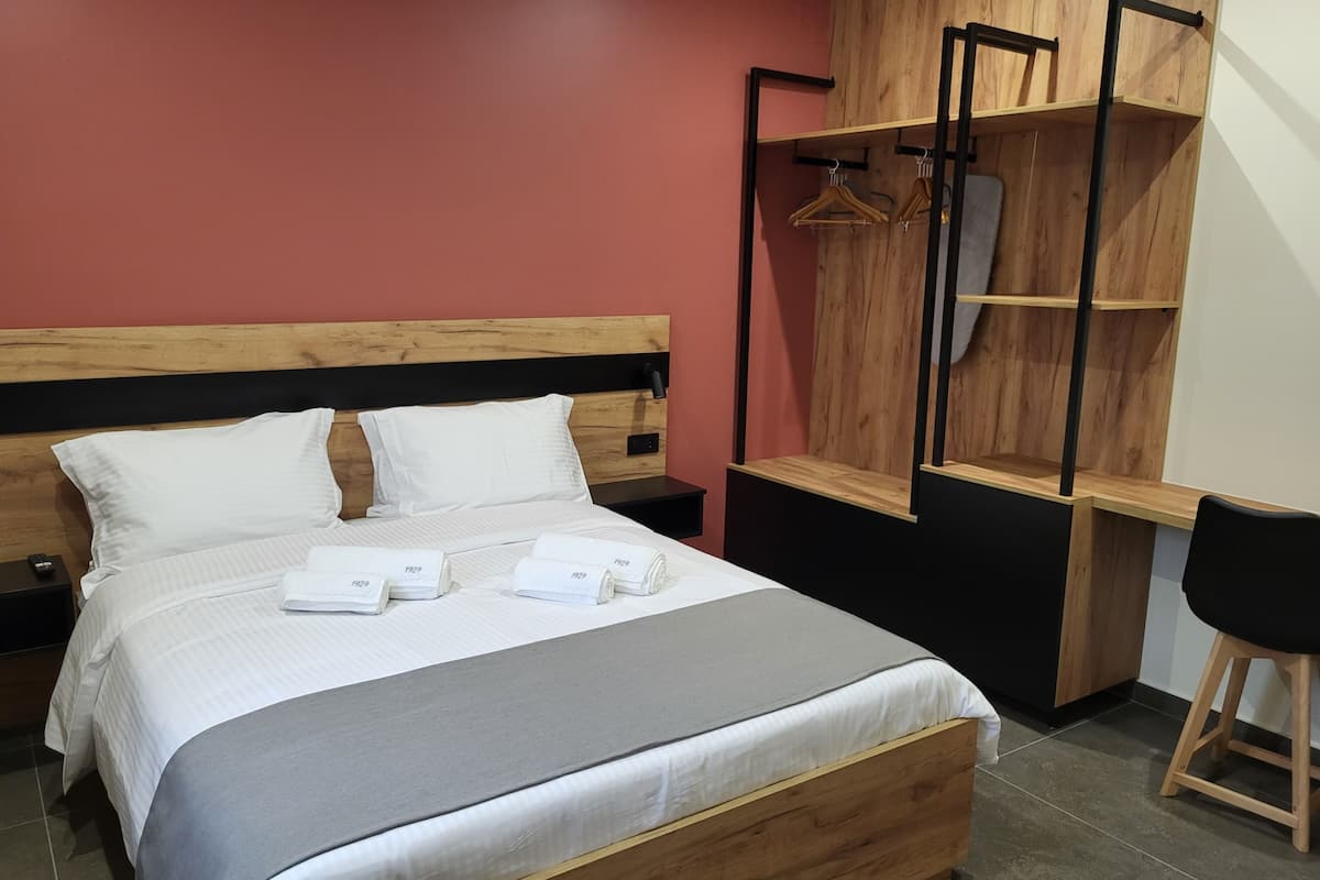 Un lit bien fait, avec du linge de lit blanc et une couverture grise, une tête de lit en bois, un bureau en bois avec une chaise et une armoire avec des étagères en bois et un miroir.