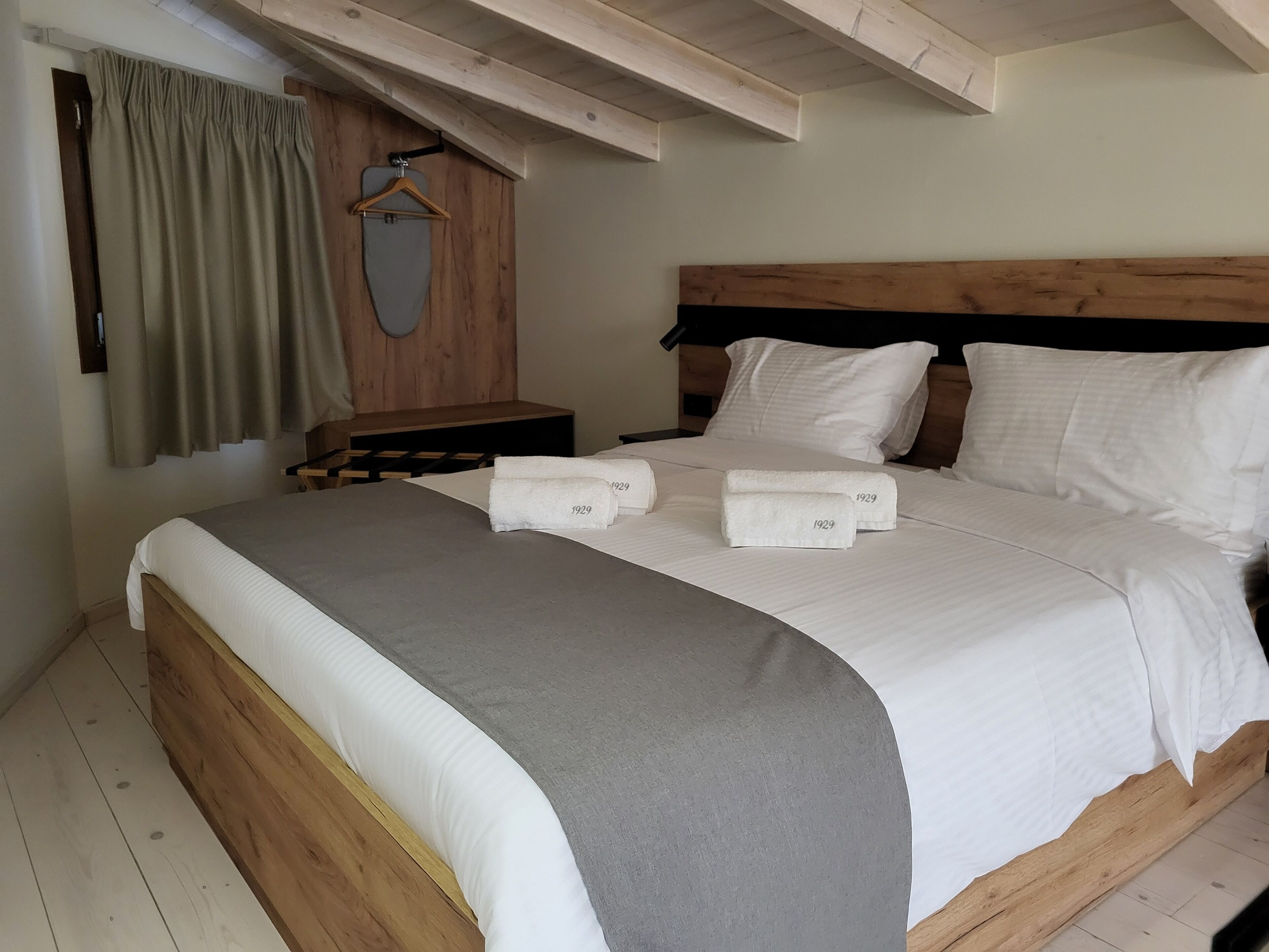 A neatly made bed with a grey blanket, two white pillows, and two rolled towels with '1929' printed on them.
