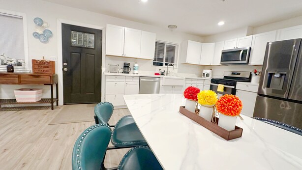 Una cocina moderna con armarios blancos, un refrigerador de acero inoxidable, un microondas y un lavavajillas. Hay una mesa de comedor con sillas color turquesa y un florero con flores.